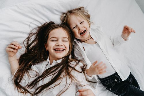 Free Two happy sisters with long hair laughing together while lying on a bed. Stock Photo