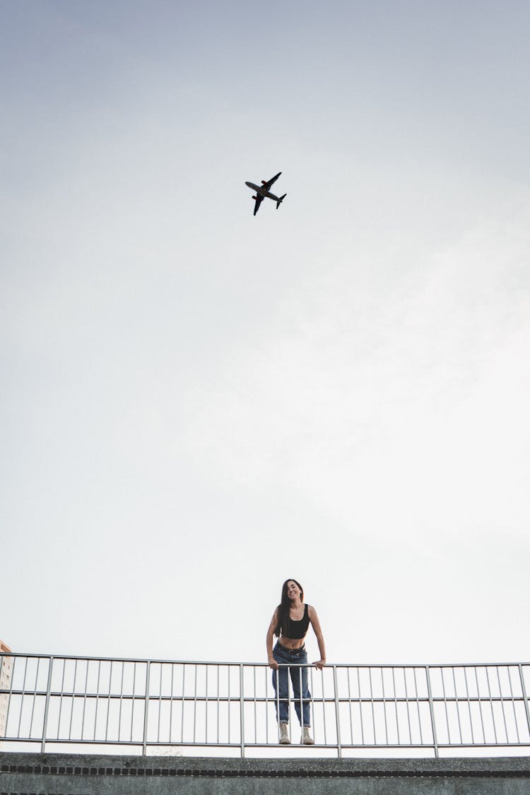 Low-Angle Shot Of Woman Standing On Metal Railings While An Airplane Flying In The Sky