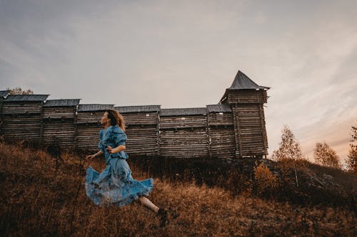 Woman in a Blue Dress Running on Brown Grass Near a Wooden House
