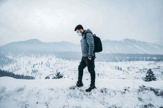 A man wearing a mask, jacket, and backpack, stands in a snowy mountainous area.