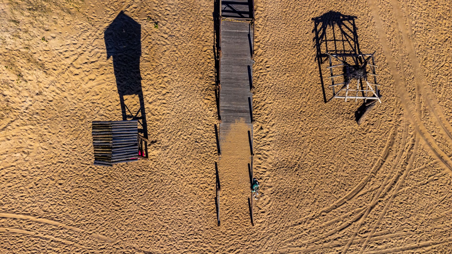 Drone shot of a wooden boardwalk and structures casting shadows on a sandy beach in Brazil.
