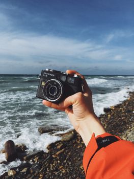Person holding a vintage camera at a rocky beach with a view of the waves.