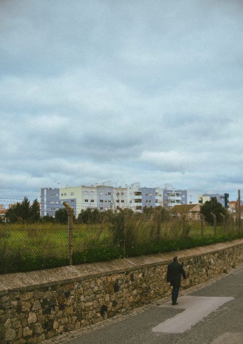 Free A man walking next to a wired fence with buildings in the background on an overcast day. Stock Photo