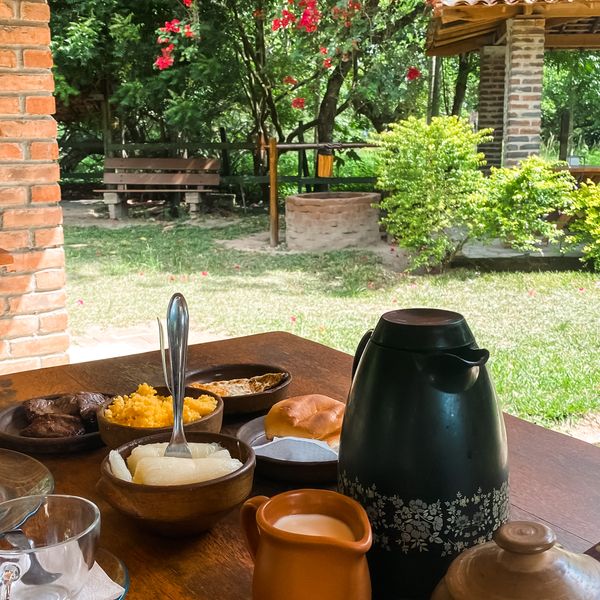 Mesa de café da manhã num alpendre, com garrafa térmica, bule de barro e jardim ao fundo