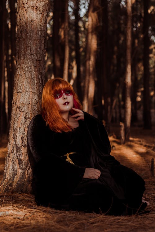 Redhead Woman with Red Painted Face Sitting under a Tree on a Brown Forest Floor