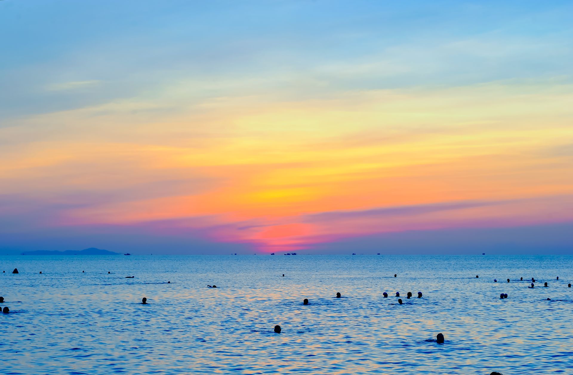 Colorful sunset over a tropical ocean with silhouettes of people swimming, creating a serene beach scene.