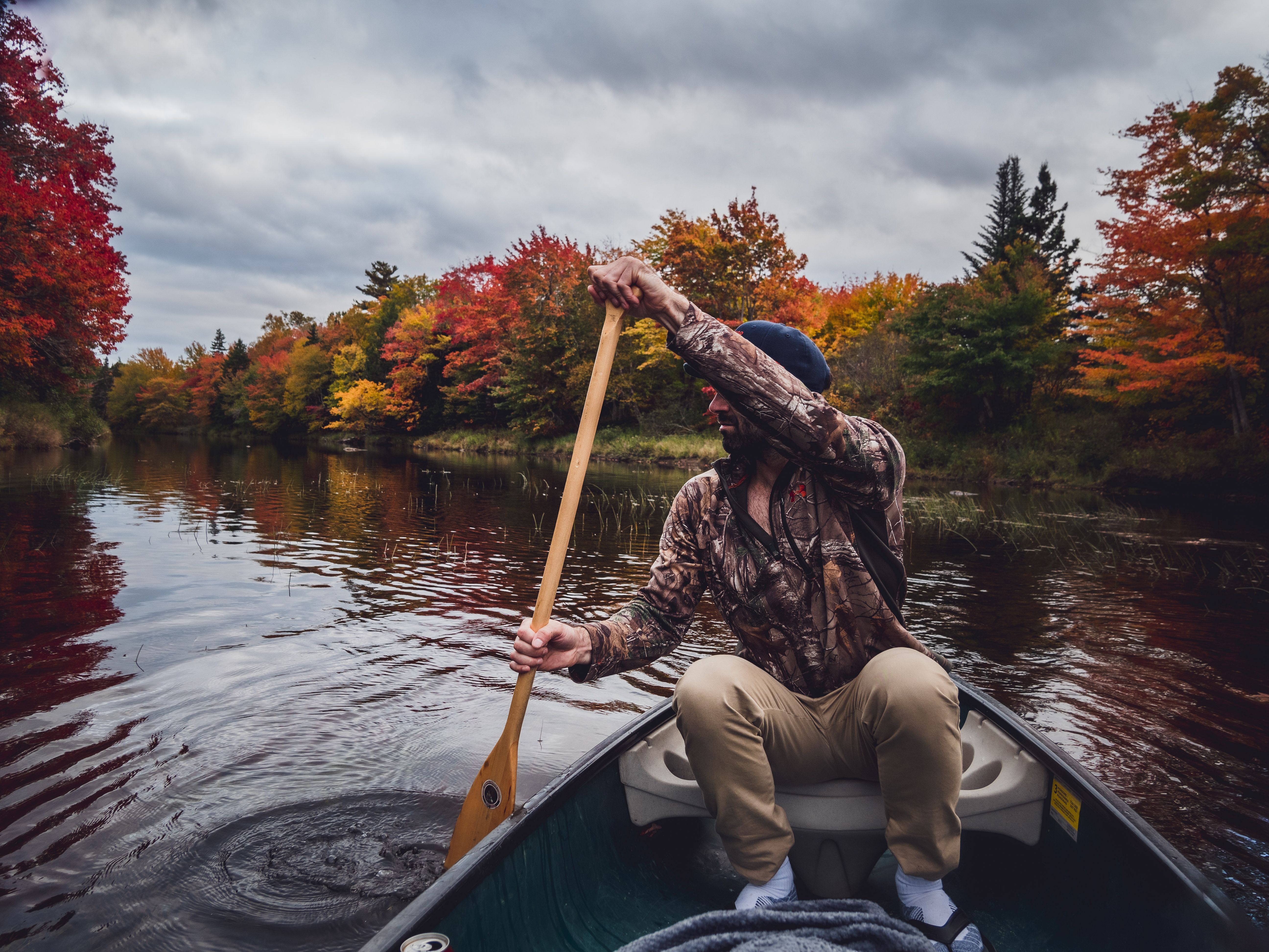 Man paddling a canoe on a calm river surrounded by vibrant fall foliage.