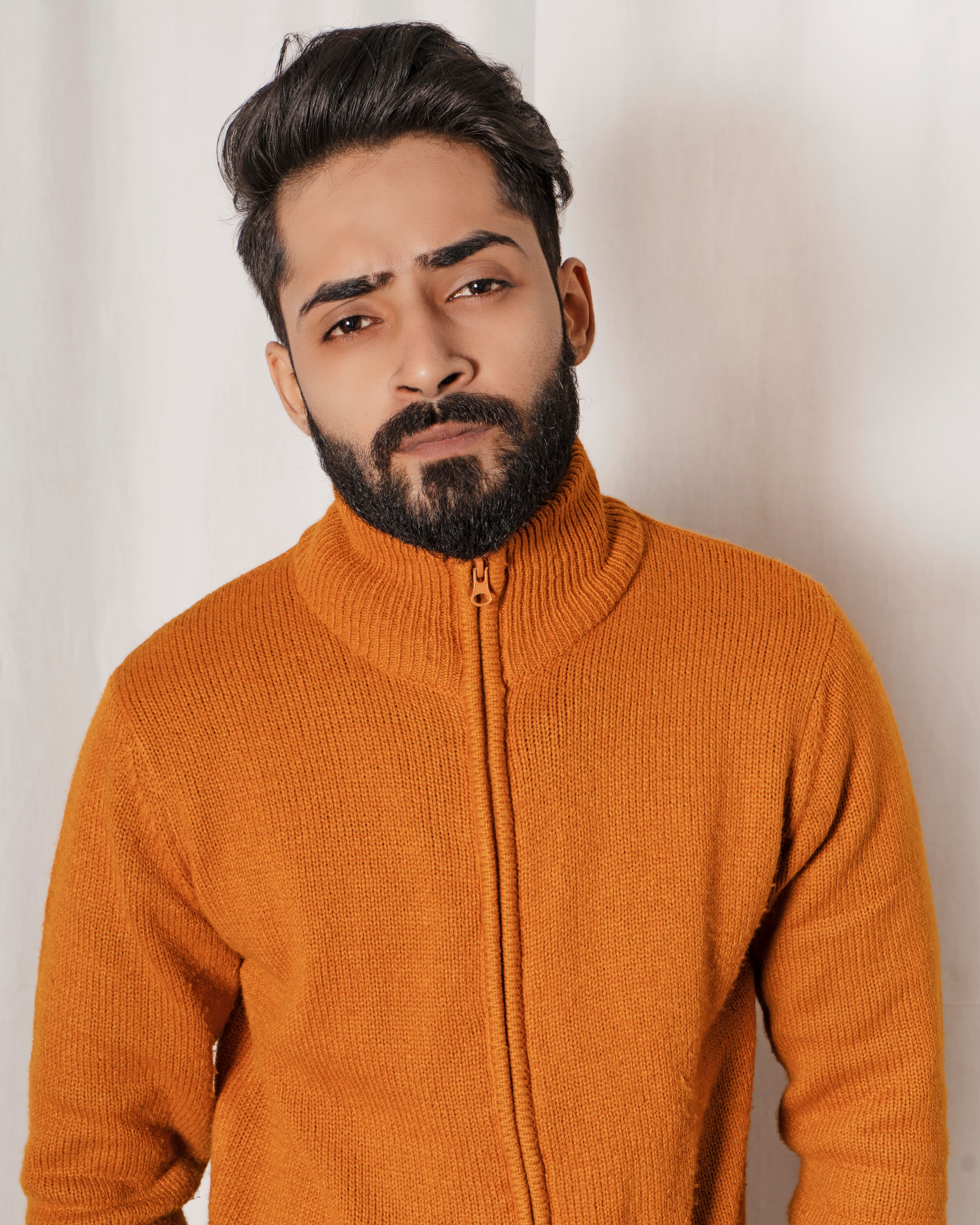 Close-up of a bearded man wearing an orange knitted sweater, looking confident indoors.