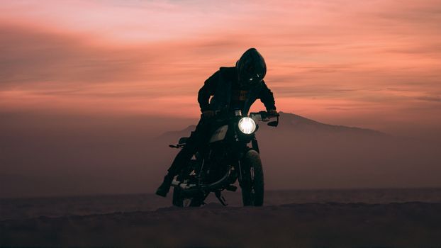 A motorcyclist rides through the desert at sunset, creating a dramatic silhouette against the orange sky.