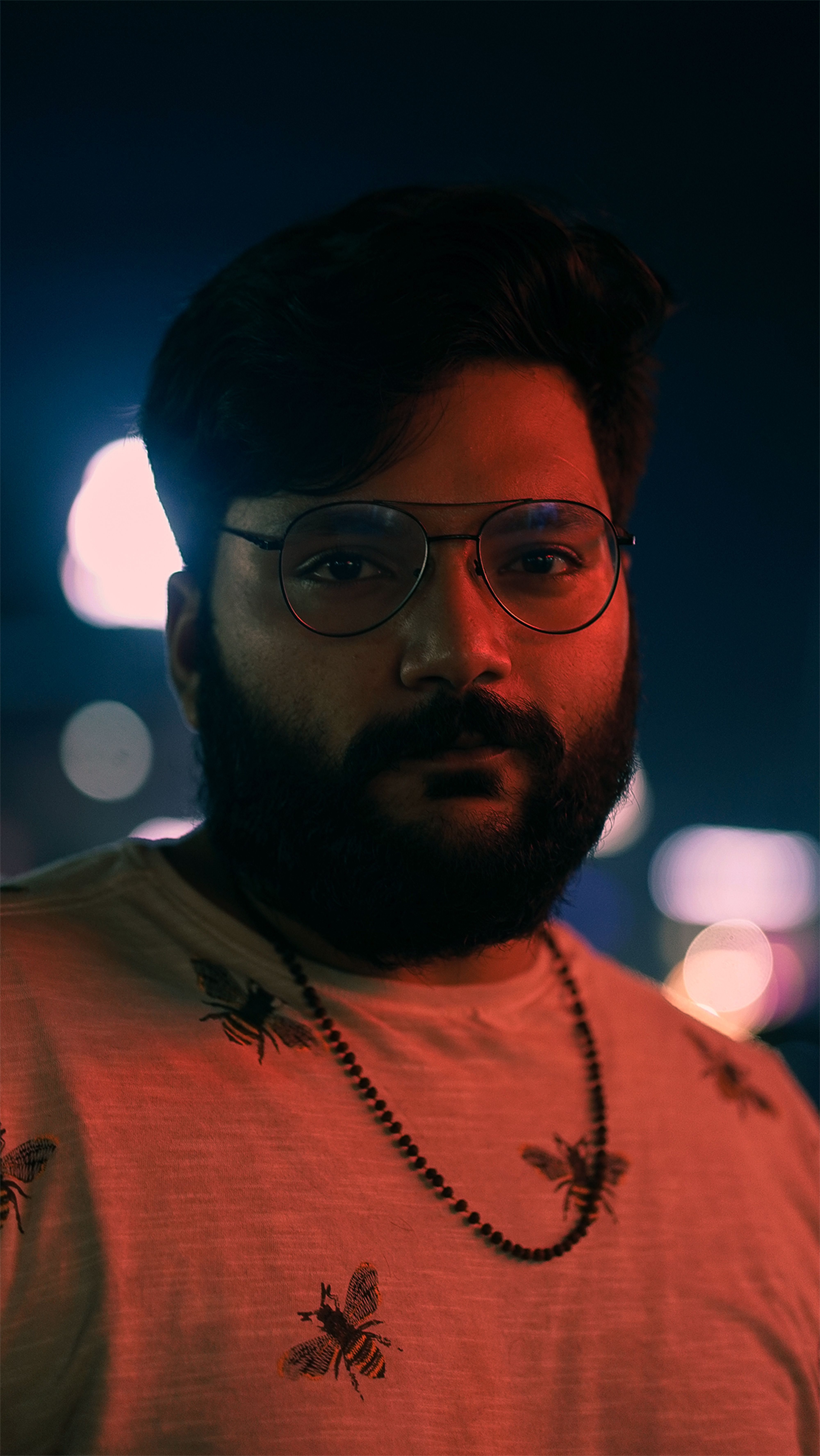 A dramatic nighttime portrait of a bearded man wearing eyeglasses, set against a blurred cityscape.