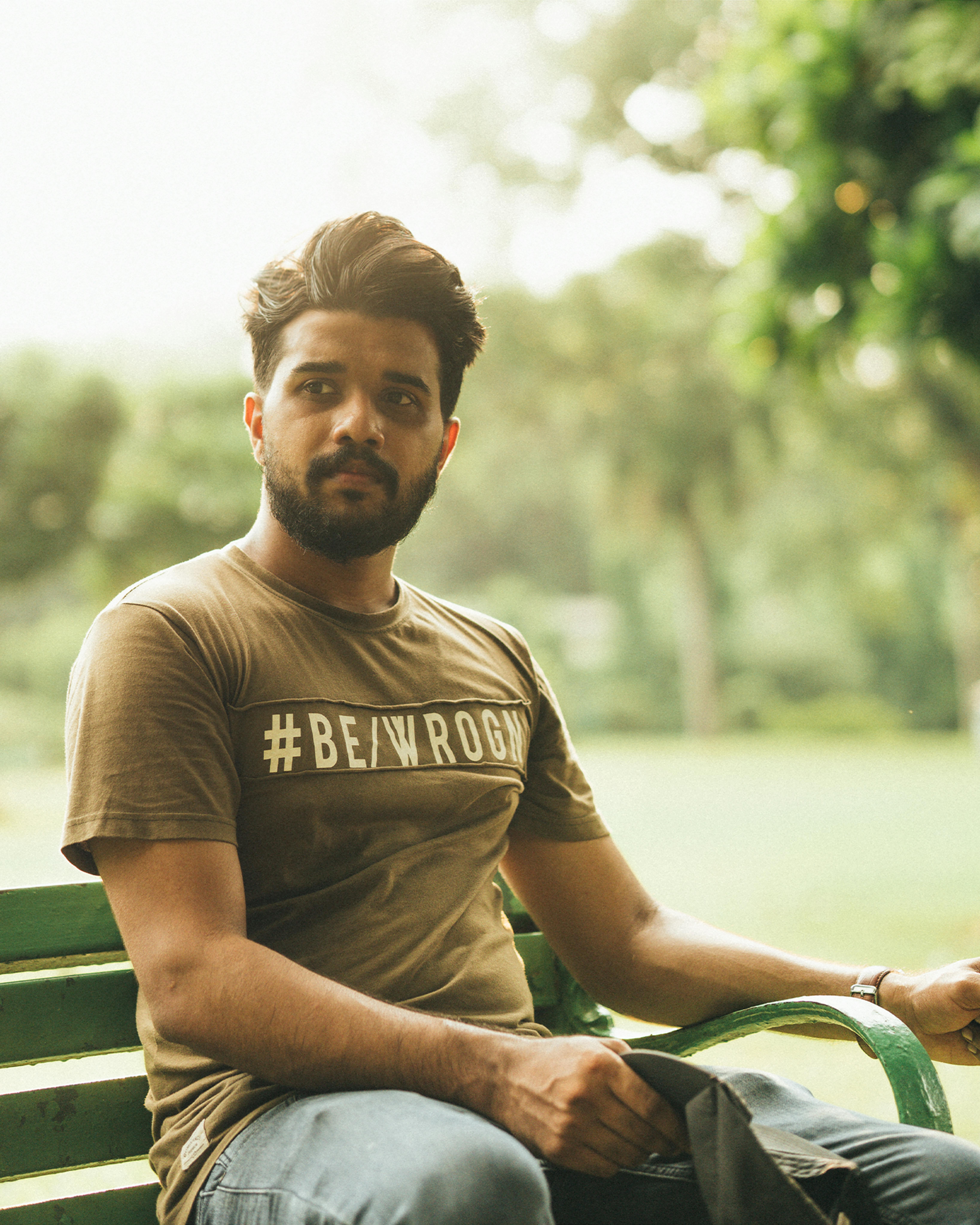 Man with beard sitting on a bench in a lush park, wearing a hashtag T-shirt.