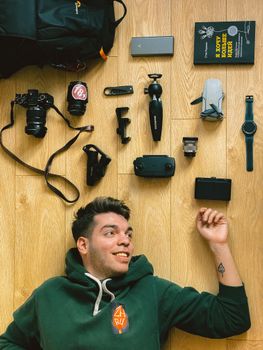 Smiling man lying on wooden floor surrounded by photography and tech gadgets.