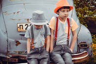 两个时尚的孩子戴着帽子和吊带，坐在户外的旧车上，显得可爱又时髦
