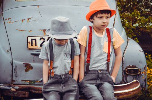 Free Two stylish children sitting on an old car outdoors, showcasing cute fashion with hats and suspenders. Stock Photo
