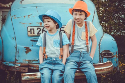 Free Two young boys in colorful hats and suspenders sit on a vintage car, enjoying a day outdoors. Stock Photo