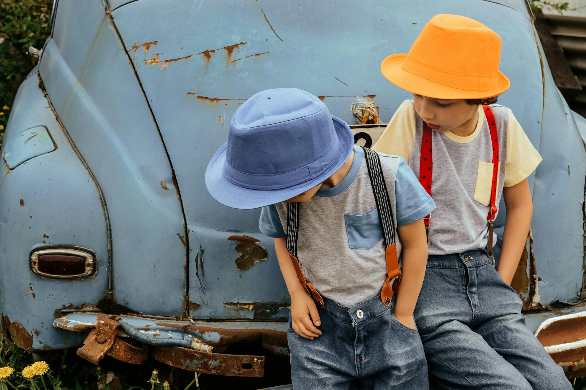 Two children in colorful hats leaning on an old rusty car outdoors.