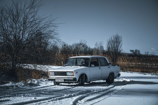 A classic white car drives on a snow-covered road in a serene winter setting, surrounded by bare trees.