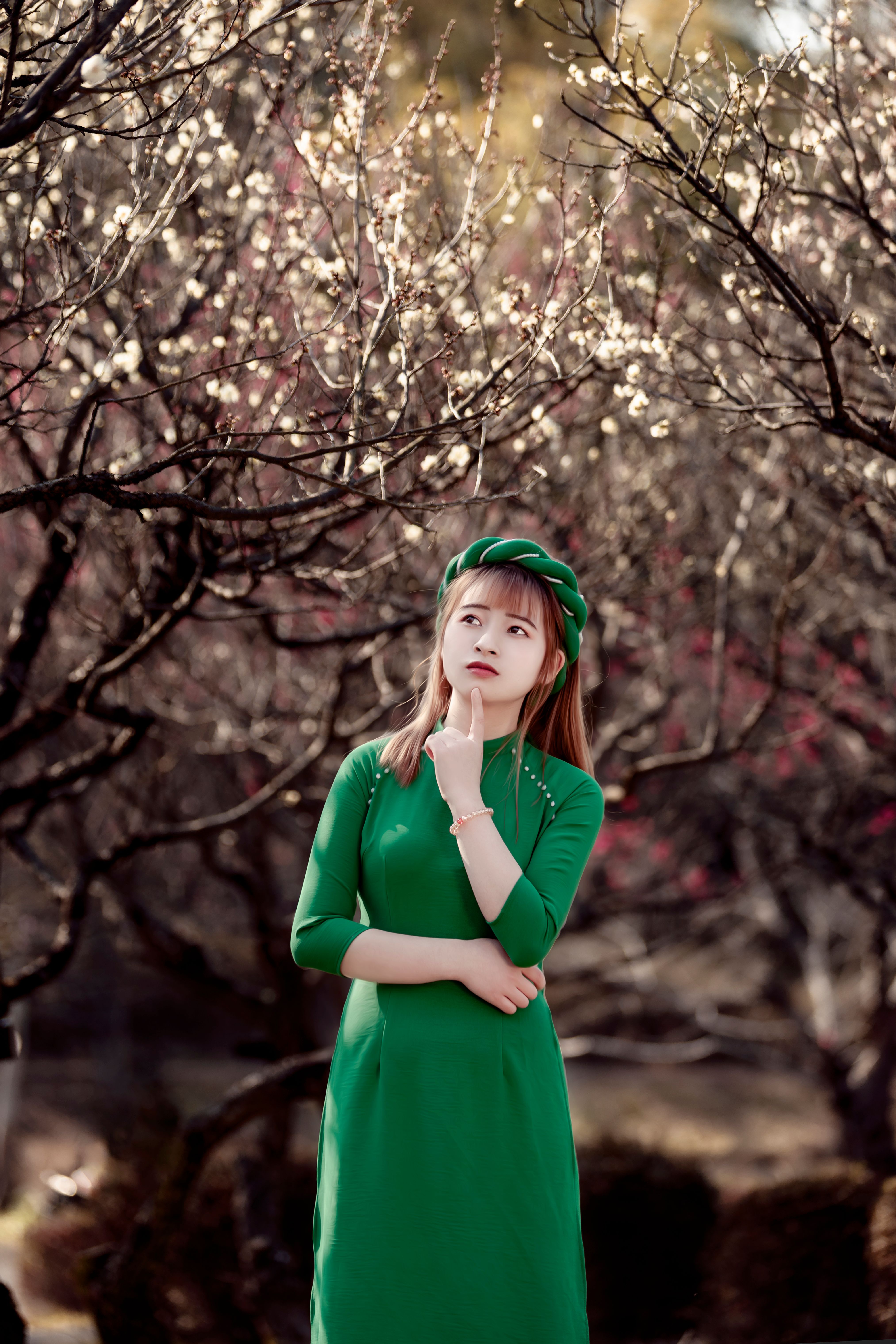A thoughtful woman in a green dress stands amidst blooming trees in an orchard, reflecting the essence of spring.