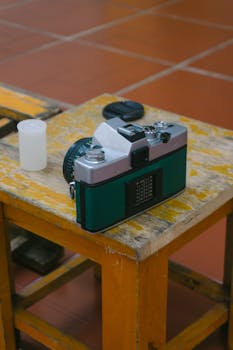 A vintage film camera rests on a worn wooden table with a film roll canister indoors.