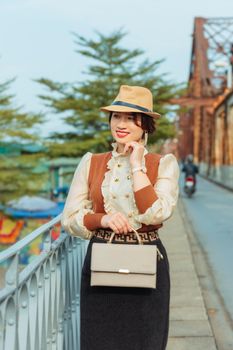 Chic woman in stylish attire standing on a bridge, holding a bag, exuding confidence.
