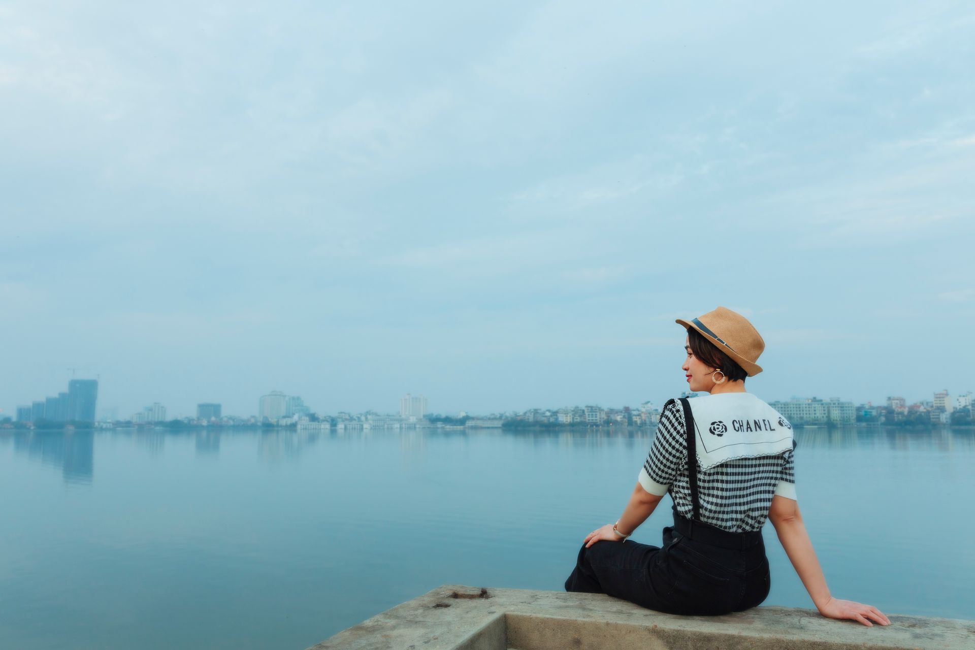 A woman in a hat sits by the river, gazing at the city skyline under a blue sky.