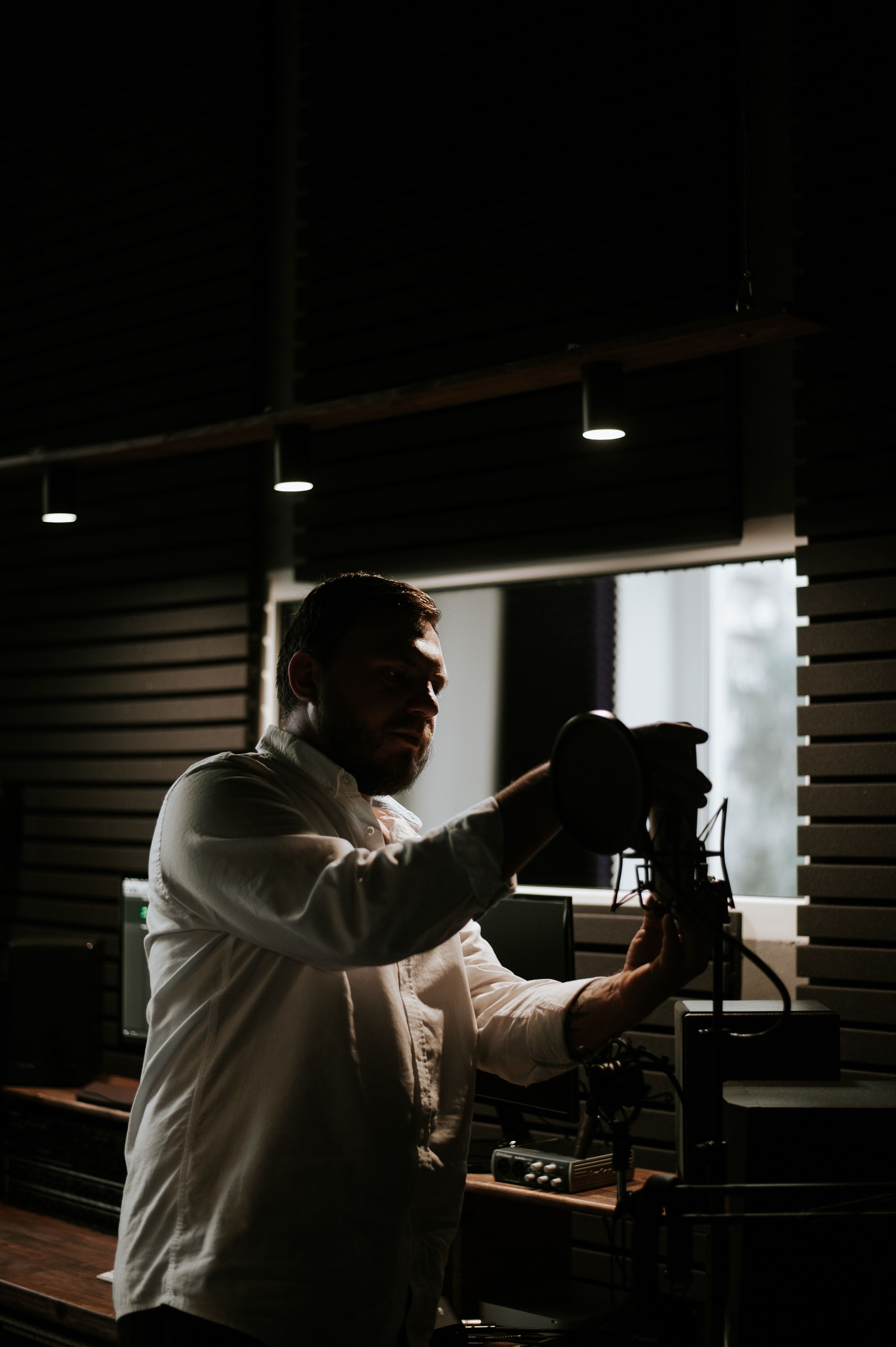 Man adjusting microphone in dimly lit recording studio, silhouetted against window.