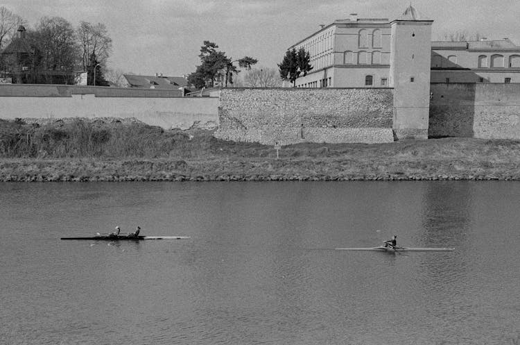 People Kayaking On River Beside Stone Wall And Building