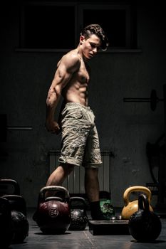Athletic man flexing near kettlebells in moody gym lighting.