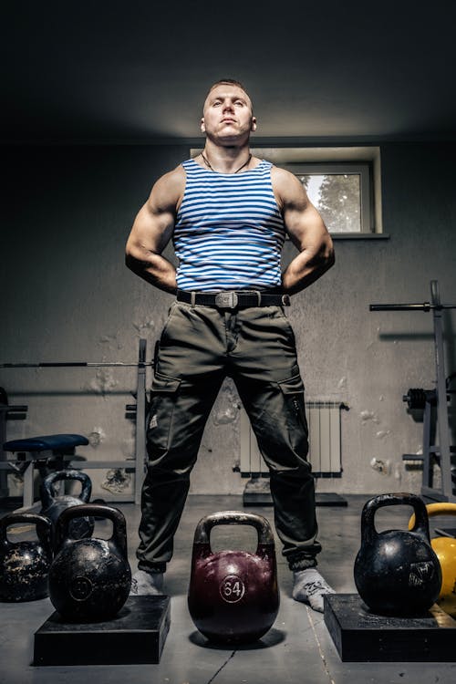 Free Man in Striped T-Shirt Standing With Hands Behind Back Beside Dumbbells Stock Photo