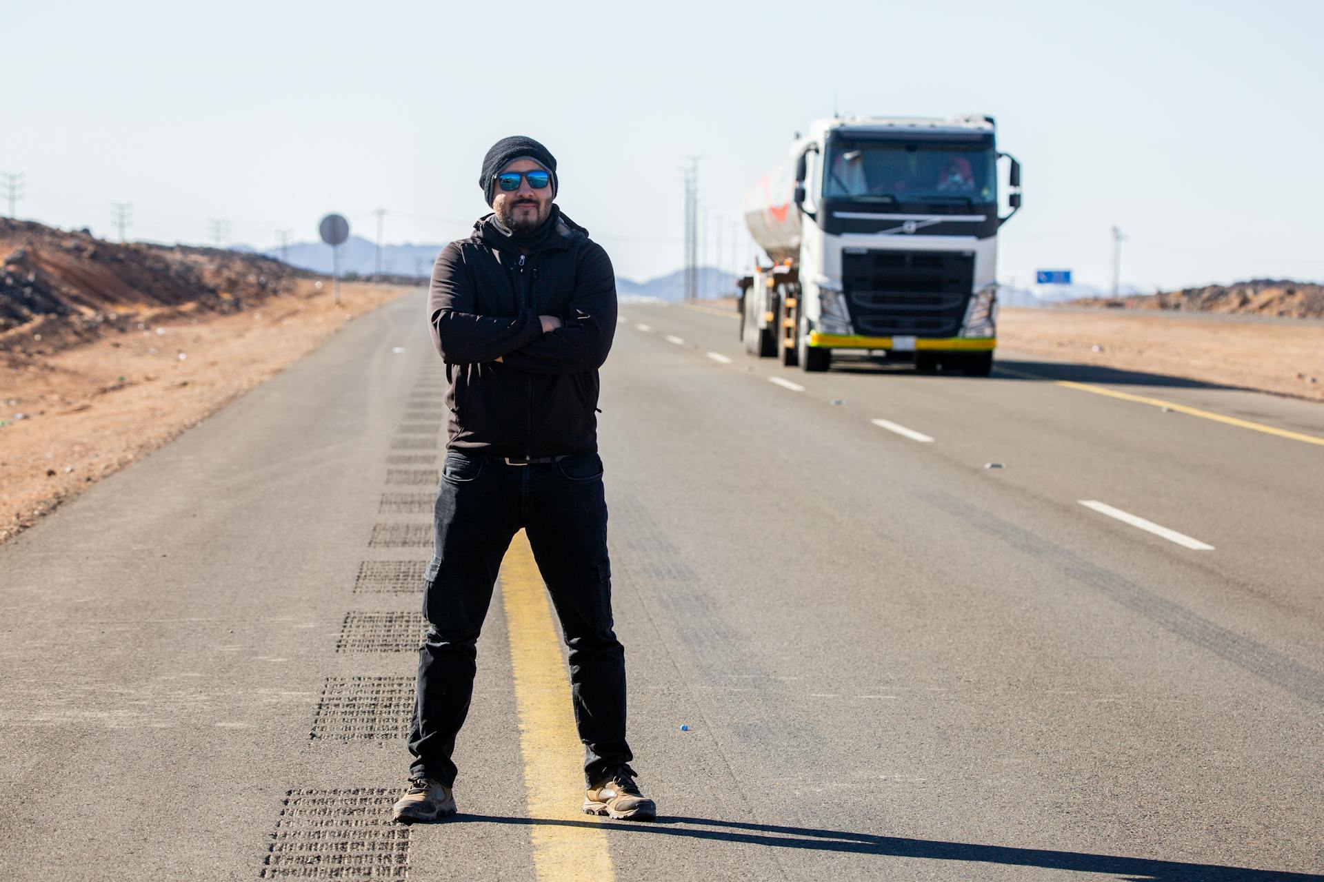 Man with sunglasses confidently stands on a desert road with a truck approaching.