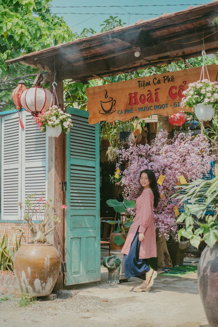 Girl In Long Pink Shirt And Flip Flops Standing Under Wooden Café Sign Beside Teal Gates