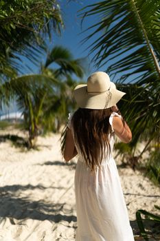 A woman in a sun hat and white dress walks on a sunny tropical beach.
