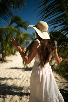 Elegant woman in white dress and hat strolls through Playa del Carmen's palm-lined beach.
