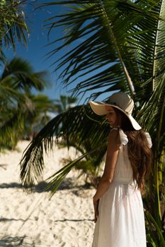 A woman in a white dress and sun hat strolls along a palm-lined beach in Playa del Carmen, Mexico.