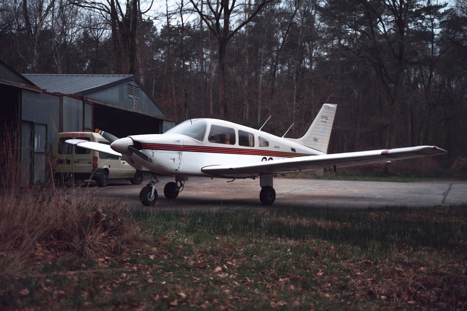 White private aircraft parked on a tree-lined airstrip beside a metal hangar amid a forest setting.