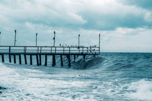Free Tranquil view of the sea with waves crashing against a pier, under a cloudy sky with seagulls perched atop. Stock Photo