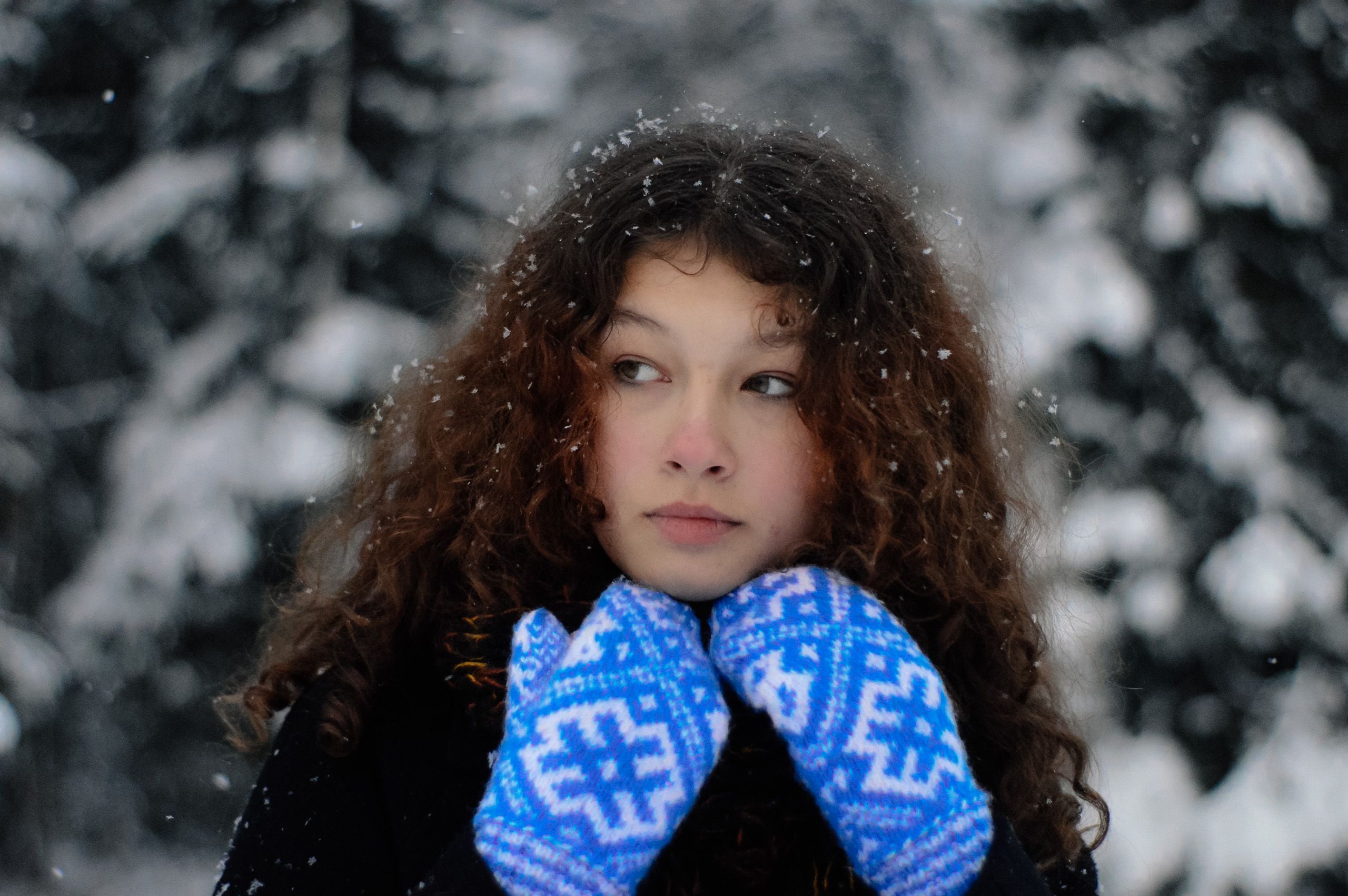 Woman with curly hair in winter attire, surrounded by falling snow in a serene forest.
