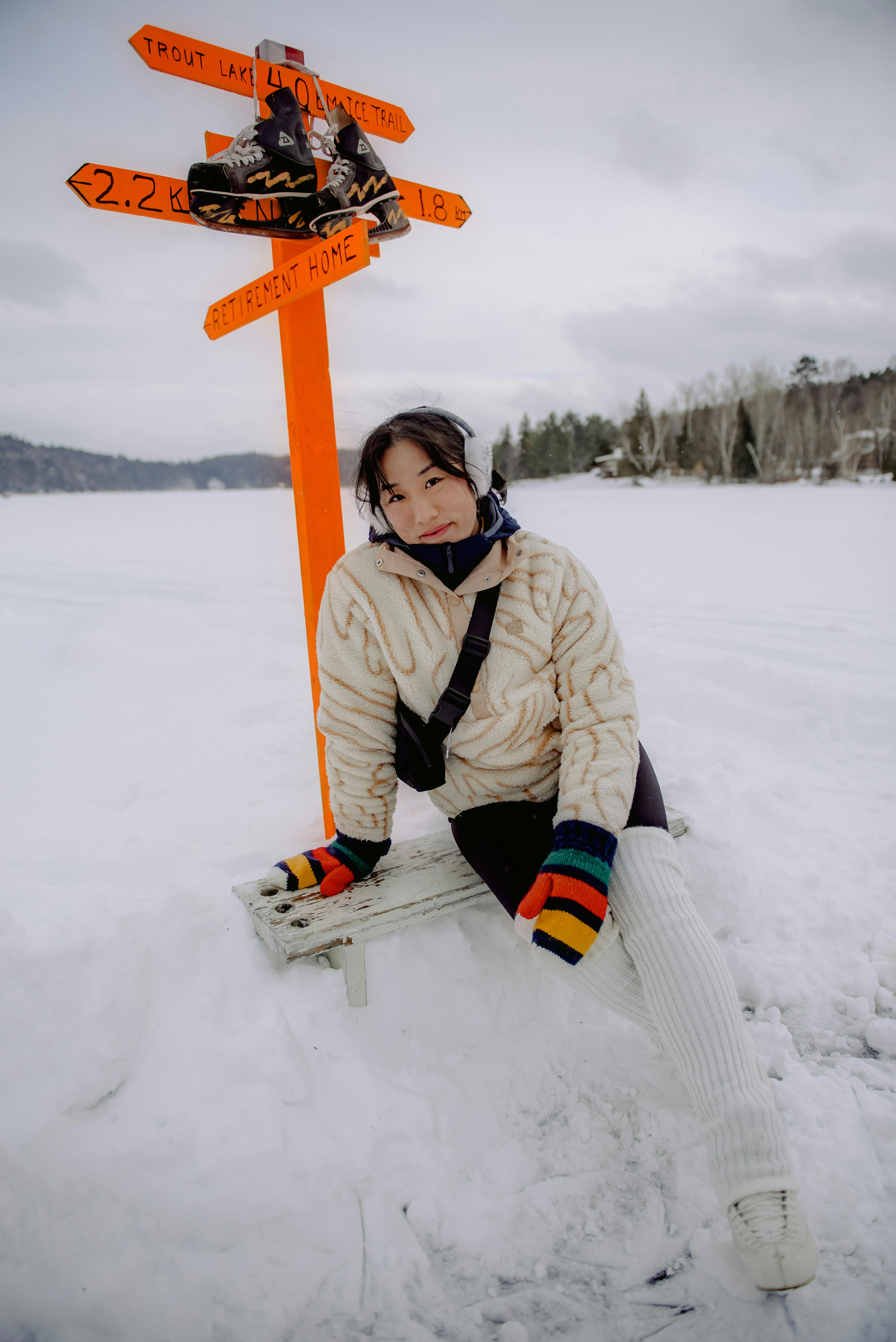 Woman sitting in winter attire near a directional sign in snowy landscape.