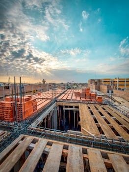 Bird's-eye view of an industrial construction site with stacks of red bricks and wooden planks under a blue sky at sunrise.
