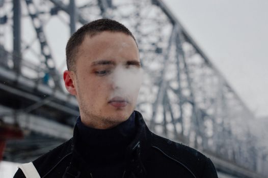 A young man exhales steam on a cold winter day, standing near a large iron bridge.