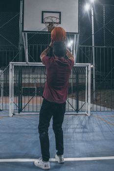 A man in casual wear shoots a basketball at an outdoor court hoop under night lights.
