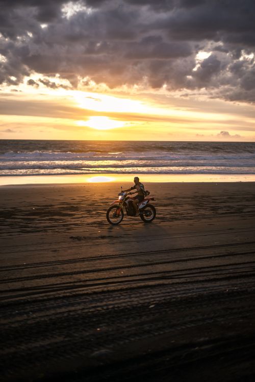 Free A motorcyclist rides along the beach with the ocean and a stunning sunset backdrop. Stock Photo