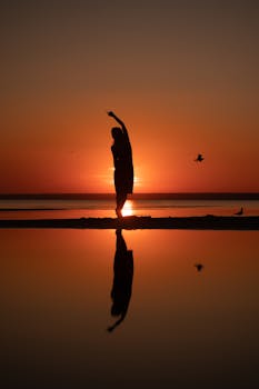 A lone silhouette stands with reflection on a beach at sunset, creating a serene scene.