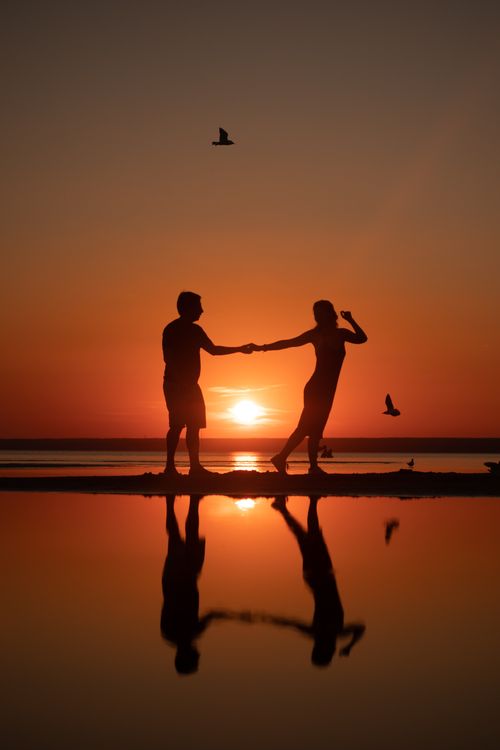 Free A couple holding hands during a stunning sunset, reflected in calm water. Stock Photo