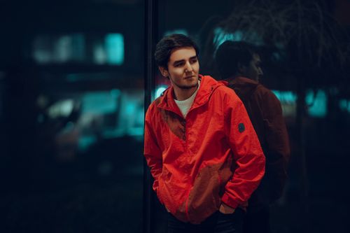 Free Portrait of a confident man in a red jacket leaning by a glass window at night. Stock Photo