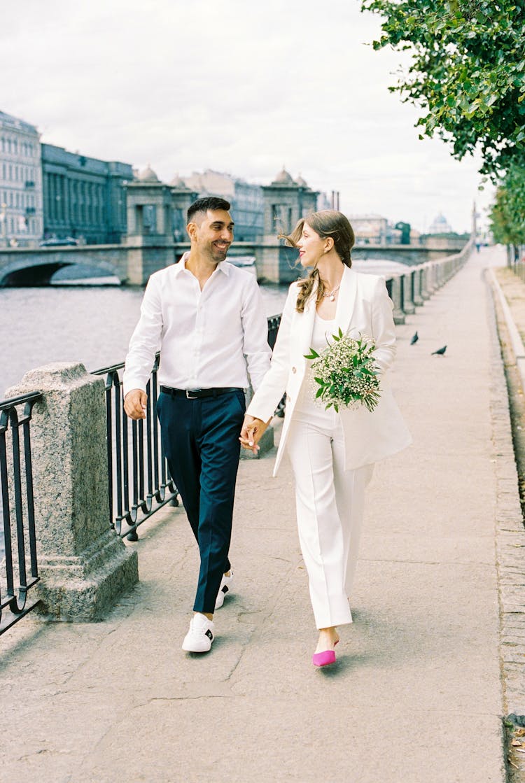 Young Couple Holding Hands And Walking Along Promenade On River