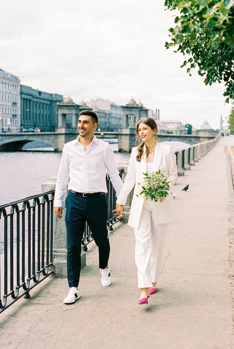 Man And Woman Holding Hands And Walking Along River With Town Bridge And Buildings In Background 