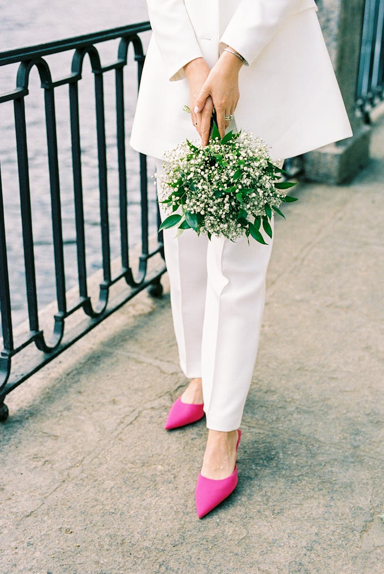 Unrecognizable Woman Wearing White Suit And Pink High Heels Standing On City Embankment With Bunch Of Flowers