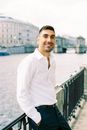 Bearded Man in White Shirt Leaning Against Railing of River Promenade and Smiling
