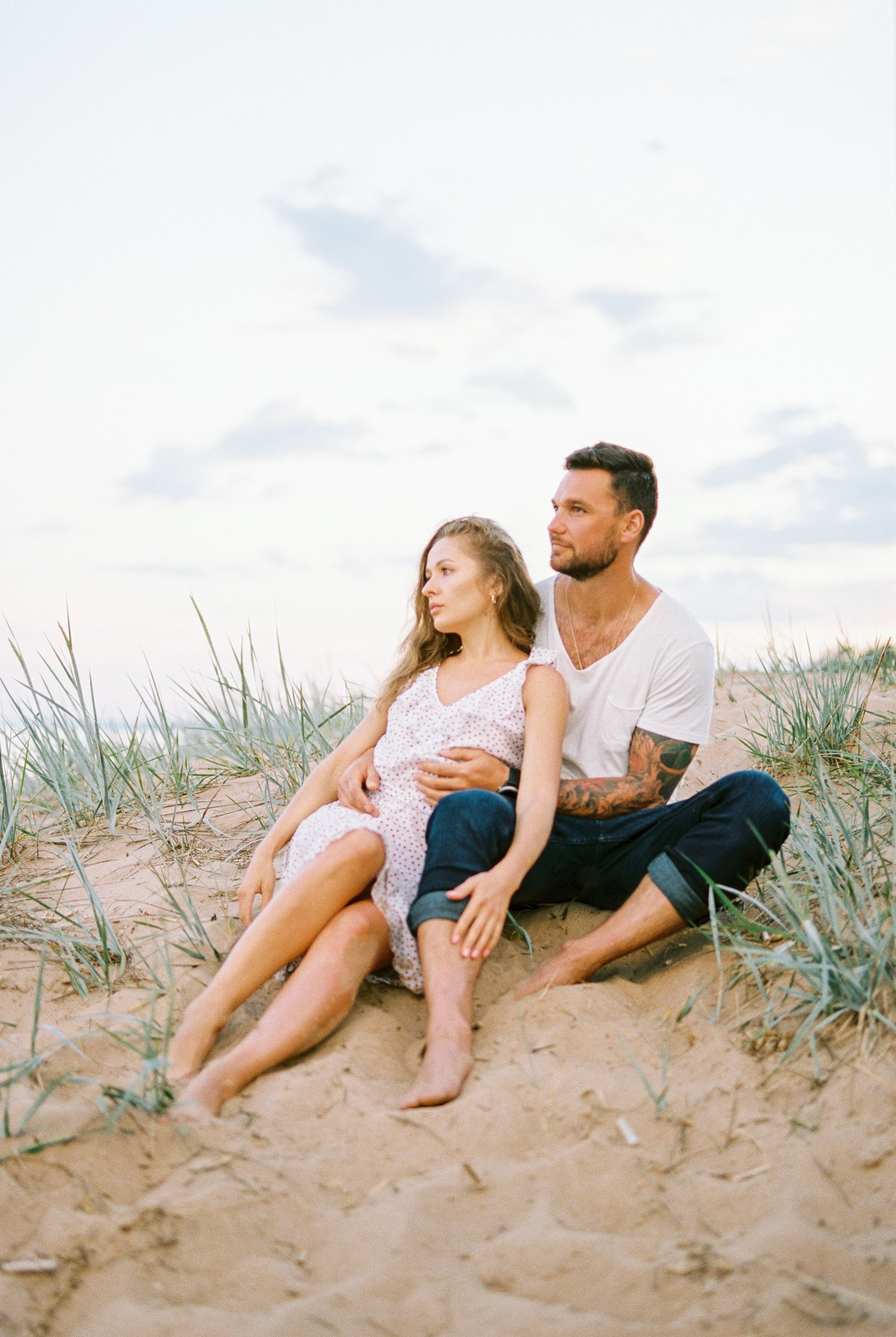 A couple enjoys a peaceful moment sitting on a sandy beach with grass dunes.